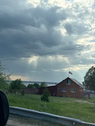 The image depicts a rural or semi-rural scene in Kryukovo, Russia, under a cloudy sky. A brick house with a Russian flag flying from its roof is visible. The house has multiple windows and is surrounded by a green lawn and trees. A fence borders the property. A utility pole with a wire is in the foreground on the left. Part of a guardrail is on the right.