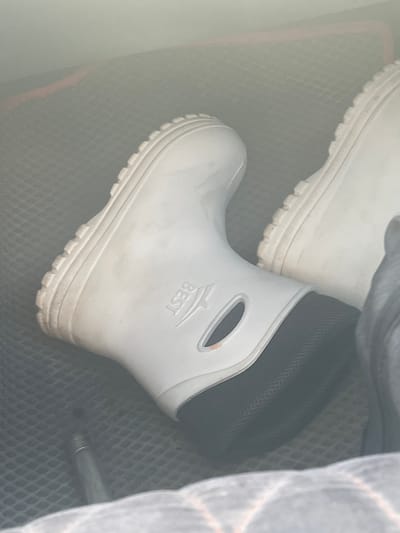 The image captures two white, short boots, likely made of rubber or PVC, stored within what appears to be a vehicle's cargo space. The boot in the foreground is prominently displayed, showing its robust, ridged sole and a black, mesh-textured lining around the ankle opening. The word "BEST" is embossed on the outer side of this boot. A second identical boot is partially visible in the upper left corner, showcasing its matching sole.
The boots are resting on a dark-colored, textured surface, possibly a rubber mat with a diamond or hexagonal pattern, typical of car trunks or floor liners. To the right, a crumpled piece of light-colored fabric, possibly a blanket or tarp, with faint reddish-grey plaid lines, partially obscures the view. A slender metallic rod or pipe is also visible near the bottom right corner of the frame.
The lighting is soft and diffused, suggesting an indoor setting or overcast conditions, and there is no indication of the time of day or specific weather. The scene depicts the boots in a state of storage, with no active event occurring. The image focuses on the objects and their immediate surroundings, conveying a practical, utilitarian context, possibly within a vehicle in Russkie Kirmeni, Russia.