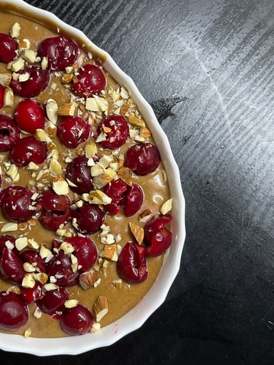 A close-up, high-angle shot shows a white oval baking dish filled with a tan-colored batter, topped with halved cherries and chopped almonds. The dish is placed on a dark, textured surface that appears to be a countertop or table. The cherries are a deep red, some showing their pits. The almonds are scattered throughout, some whole, some broken into pieces. The batter appears smooth and slightly glossy. The scene is likely indoors, possibly a kitchen, in Калодзішчанскі сельскі Савет, Belarus, though no specific location markers are visible.