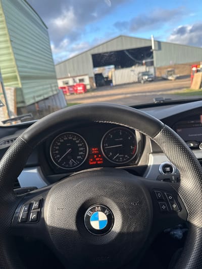 The image depicts the interior of a BMW vehicle, specifically the steering wheel and instrument cluster, from the driver's perspective, looking out onto an industrial or agricultural yard in Fowlis Easter, United Kingdom.
In the immediate foreground, a black leather-wrapped steering wheel, featuring a centered BMW logo with blue and white quadrants, dominates the lower half of the frame. The steering wheel has perforated leather on the right side and integrated control buttons on both spokes: volume and phone controls on the left, and four diamond-shaped function buttons on the right. Behind the steering wheel, the vehicle's instrument cluster is visible, displaying a speedometer on the left and a tachometer on the right, both indicating zero. The central digital display shows the time as 16:41, an outside temperature of +7.0°C, a remaining fuel range of 537 km, and an odometer reading of 250787.0 km. A 'START STOP' engine button is visible on the dashboard to the right of the steering wheel. Sunlight casts a defined shadow across the steering wheel and dashboard from the upper left.
Through the windshield, a rural or semi-industrial outdoor environment is visible under a partly cloudy sky with patches of blue. To the left, a large, light-green corrugated metal structure, possibly a mobile unit or temporary building, stands partially visible. In the background, a substantial, light-grey corrugated metal shed or barn with a wide, dark, open entrance bay forms the primary backdrop. In front of this main building, a smaller, white building with windows, two red skip bins, a large white corrugated shipping container, a dark-colored sedan parked behind the container, and a small trailer are discernible. The ground consists of dirt and gravel. No individuals are visible in the scene. The vehicle is stationary with its engine either running or accessory power engaged.