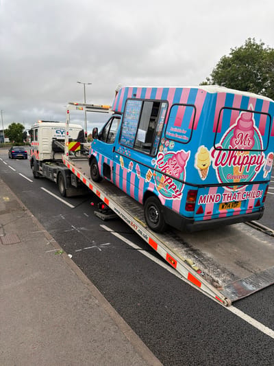 The image depicts an ice cream van, branded "Mr Whippy Gloucester," being loaded onto a flatbed tow truck. The van is predominantly blue and pink with vertical stripes and features various ice cream illustrations and the slogan "MIND THAT CHILD!". The tow truck, labeled "COTSWOLD CVI," has an orange and white striped beacon light bar on its cabin and "RESCUE247.CO.UK" printed on its ramp. The scene appears to be on a road with white road markings and a paved sidewalk. The sky is overcast. A dark blue car is visible in the background on the road. The overall impression is that of a vehicle recovery operation.