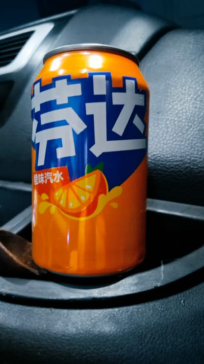 A close-up, low-angle shot captures an orange soda can with prominent blue and white Chinese characters, "劳达," on its side. A sliced orange with a splash of juice is depicted on the lower portion of the can, suggesting an orange flavor. The can rests in a dark holder, likely within a vehicle, as indicated by the textured dark grey material and a portion of a silver vent or console in the blurred background. The lighting is dim, creating a slightly moody atmosphere. The location is not specified within the image itself.