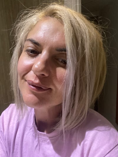 The image is a close-up selfie of a woman with light blonde hair and brown eyes. She is wearing a light purple t-shirt. The background is dark and out of focus, suggesting an indoor location. The woman is looking at the camera with a slight smile. There is no evidence to suggest that the image was taken in Monastir, Tunisia.