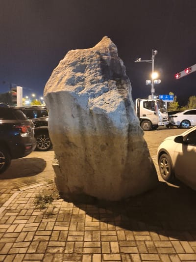 The image depicts an outdoor scene at night, illuminated by streetlights and traffic signals. The foreground features a large, rough-textured boulder casting a prominent shadow on a paved area constructed with interlocking bricks. To the left of the boulder, the rear of a dark-colored SUV is visible. Behind the boulder, to the right, a white truck is parked on a road. Further to the right, a white sedan is parked. In the background, a traffic light with red illumination is visible, along with street lamps and trees. A blue street sign with Chinese characters is also present, indicating the location in Jiguan District, China. The scene appears to be a public space, possibly a roadside or a parking area. There are no people visible in the image.