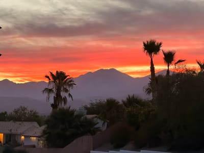 A dusk sky over Desert Hot Springs, California, features a vibrant gradient of orange, red, and purple hues. The sky is filled with wispy clouds, with the brightest colors concentrated near the horizon. Silhouetted palm trees and other foliage are visible in the foreground, along with the roofs of several houses. In the distance, mountains form a hazy backdrop against the colorful sky. The overall scene depicts a serene desert sunset.