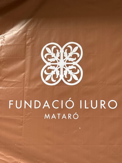 The image shows a white logo on a brown, wrinkled surface. The logo consists of a stylized floral or quatrefoil design at the top, and text below. The text reads "FUNDACIÓ ILURO" and, centered below that, "MATARÓ". The surface appears to be a banner or sign. The location is confirmed by the text "MATARÓ", which is the name of a city in Catalonia, Spain.