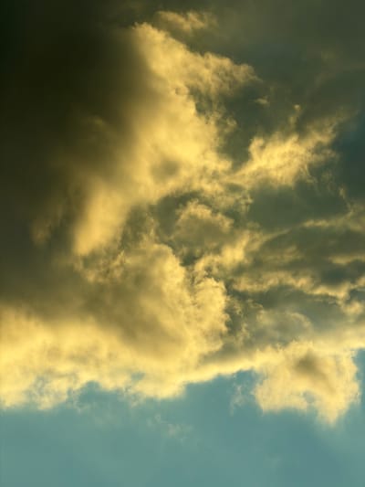 The image is a vertical, eye-level, close-up shot of dramatic, turbulent clouds against a teal-colored sky. The clouds are lit from the left, with bright yellow and golden hues highlighting their fluffy textures, while the right side of the image is darker with deep grays and blacks. A band of lighter, more wispy clouds separates the upper, darker clouds from the clearer sky below. The scene appears to be an outdoor sky view, possibly taken in Studena, Bulgaria, given the context provided. There are no people, subjects, objects, or actions depicted.