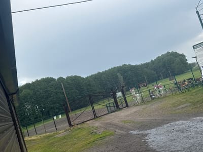 The image depicts an entrance to an outdoor recreational area or park in Zaliznychne, Ukraine. In the foreground, a dirt path with tire tracks leads towards a large, ornate black metal gate. To the left of the path is a portion of a dark, corrugated building. Beyond the gate, a fenced-off grassy area is visible, with a dense line of trees forming the background. Decorative elements, including a white metal bicycle with flower baskets and a white metal reindeer figure, are present near the gate. In the distance, tables and benches suggest an outdoor seating area. A sign with text is attached to the fence near the gate, and another sign with Ukrainian text is visible on the gate itself. The sky is overcast.