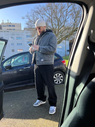 The image depicts an outdoor urban scene in Hérouville-Saint-Clair, France, observed from inside a parked vehicle. A male subject, appearing to be in his 20s or 30s with a dark beard, stands on a paved surface. He wears a white knit beanie, light-tinted sunglasses, a faded grey quilted jacket over a black hooded sweatshirt, black loose-fitting trousers, and grey sneakers with white accents. The man is holding a black smartphone horizontally with both hands, focusing on its screen. A plume of white smoke or vapor is visible originating near his right hand. Behind the subject, a dark-colored hatchback vehicle, possibly a Citroën C3, is parked with its front passenger door open. In the background, two multi-story white residential buildings are visible under a clear blue sky. A large deciduous tree with bare branches stands between the buildings and the foreground. The scene is brightly lit by direct sunlight, casting distinct shadows.