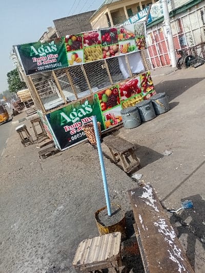 The image depicts a roadside scene in the city of Sharada, Nigeria, centered on a fruit juice stall under daylight conditions.
The primary subject is "Aga's Fruits Mix & Juices" stall, constructed from a light-colored frame (possibly wood or metal) with mesh or wire-like panels. The stall features two prominent green banners, one on the upper section and one on the lower, displaying the business name "Aga's Fruits Mix & Juices" in white text and a phone number "08036030062". Both banners are flanked by colorful photographic collages of various fruits, including grapes, pomegranates, bananas, apples, mangoes, oranges, and other berries.
In front of and around the stall, on a concrete pavement, are multiple seating arrangements: several small, simple wooden stools and a longer, weathered wooden bench with peeling white paint. To the right of the stall, three cylindrical metal containers, possibly bins or drums, are lined up on the ground. A light blue pole, anchored vertically in a concrete-filled metal bucket, stands in the mid-ground.
The immediate environment consists of a dusty, uneven concrete pavement adjacent to an asphalt road, partially visible on the left. A yellow and orange tricycle (keke napep) is partially visible on the road in the far left background. Behind the stall, a two-story building with light-colored walls and multiple windows/balconies is present. Another building further back displays text, partially readable as "AMA MINI MALI". A dark-colored bicycle is leaning against a metal fence on the right side of the image. No individuals are clearly identifiable interacting with the stall or its immediate vicinity. The sky is clear and light blue.