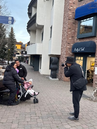A photographer in a black outfit with a camera takes a photo of a family with a stroller. The family includes two adults and a child in the stroller, all warmly dressed. They're situated on a brick-paved street near a building with storefronts, including one named "Artful Sol." A blue awning, public restroom sign, and bare tree add to the winter ambiance of the scene.