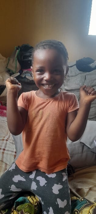 An indoor photograph, taken in the city of Ashipa, Nigeria, features a young, dark-skinned child positioned kneeling or squatting, facing the viewer directly. The child exhibits a wide, open-mouthed smile, revealing teeth. Both hands are raised to chest height, clenched into fists. The child wears an orange short-sleeved t-shirt and dark grey leggings adorned with light-colored shapes resembling the African continent. Small golden hoop earrings are worn. The background consists of a light brown wall on the left and a brightly illuminated window on the upper right. Behind the child, an assortment of objects, including dark and grey fabrics and bags, is present, indicating a cluttered interior space. The child kneels on a surface covered by a beige and brown checkered fabric, with an additional multi-colored patterned fabric visible below the child's knees.