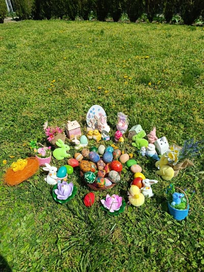 The image shows an outdoor scene on a grassy area, possibly a lawn, with various Easter-themed decorations. The decorations include a basket filled with decorated eggs, Easter bunnies, chicks, flowers, and miniature houses. The environment is likely a garden or yard, with dandelions visible in the grass. The setting is Cherkovitsa, Bulgaria. There are no people visible in the image. The overall impression is that of an Easter display or arrangement.
