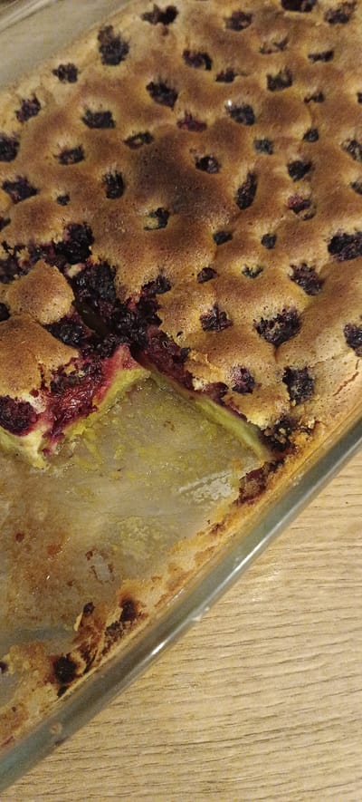 A close-up, high-angle shot shows a square glass baking dish containing a baked dessert, likely a cake or cobbler, with visible dark berries such as blackberries or blueberries embedded in a golden-brown batter. A slice has been removed from the front left corner, revealing the layered interior which includes the berries and a slightly undercooked, pale yellow layer, possibly fruit or dough. The batter has a slightly uneven, bubbly texture with some darker, crisped edges. The baking dish rests on a light-colored wooden surface with a visible wood grain. The location is stated to be Падлабенскі сельскі Савет, Belarus.