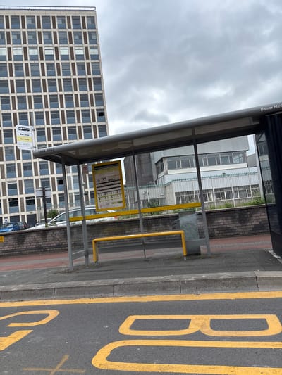The image shows a bus stop shelter with a yellow bench and timetable. The bus stop is labeled "Bus Stop Rail House". In the background, there is a tall, multi-story building with many windows. The bus stop is located on a street with yellow markings on the road, indicating a no-parking zone. The sky is cloudy. The bus stop appears to be located on the outskirts of Crewe, United Kingdom.