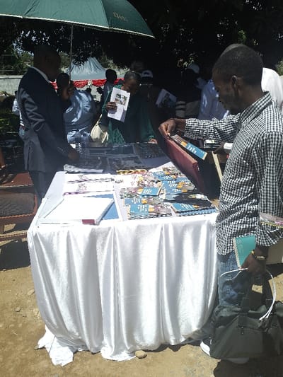 The image shows a table covered in white fabric, located outdoors on a dirt ground. The table is covered in a variety of pamphlets and brochures. A man in a plaid shirt stands by the table, reaching toward a pamphlet, and a man in a suit stands nearby. There are other people blurred in the background. The scene is likely in an outdoor market setting. The coordinates provided, latitude 1.556, longitude 30.244, place the location in the city of Bunia, Democratic Republic of the Congo. The image may show a man buying or viewing pamphlets in a market.