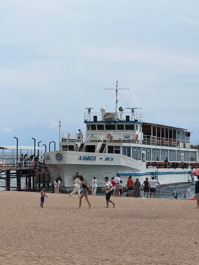 The image shows a large white passenger boat docked at a pier on a body of water, with a sandy beach in the foreground. The boat has the name "АЛЫКУЛ" and "ИК 24" written on its side. Several people are visible on the boat, on the pier, and on the beach. On the beach, a young boy in blue shorts is standing, holding a pink toy gun. Further back, a group of people are walking and standing near the water, with some individuals appearing to be playing with a ball. In the background, mountains are visible through a hazy sky. The scene is set in Bosteri, Kyrgyzstan, a resort town located on the northern shore of Issyk-Kul Lake.