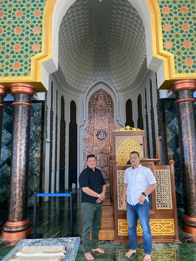Two men stand in a mosque in Aluekambuk, Indonesia. The man on the left wears a black shirt and green pants, while the man on the right wears a white patterned shirt and blue jeans. They are barefoot. Behind them is an intricately decorated mihrab and minbar, common features in a mosque. The mosque interior features marble flooring, ornate columns, and a domed ceiling with geometric patterns. A blue stanchion barrier and prayer rugs are visible on the floor. The overall ambiance suggests a place of worship with rich cultural and architectural elements.
