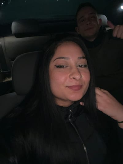 The image captures two individuals within the dark interior of a vehicle. In the foreground, a female with long, dark hair, wearing a dark puffer jacket, appears to be seated in the front passenger or driver's seat. She has a nose ring on her right nostril and winged eyeliner, and her left hand is positioned near her neck. She is looking directly at the camera with a faint smile. Behind her, in the rear seat, a male subject with short, dark hair is visible, wearing a dark hooded garment. He is looking towards the camera and displaying a 'thumbs up' gesture with his right hand. The vehicle's interior features dark upholstery, and the overall lighting is dim. The specific geographic location in Gerakas Municipal Unit, Greece, cannot be determined from the visual information present in the image.