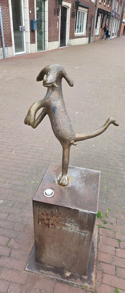 A bronze sculpture of a bipedal dog-like figure stands on a rusted metal cube pedestal on a brick-paved sidewalk in Horst, Netherlands. The sculpture, exhibiting a dark brown and greenish patina, is posed on its left leg with its right leg bent and extended backward, while its left paw is raised. A prominent penis is visible on the figure. The pedestal features engraved script on its front face and a silver-colored push-button on its top surface, to the left of the sculpture's left foot. The ground is paved with reddish-brown rectangular bricks, with minimal grass growth in the cracks. In the background, a row of multi-story brick buildings lines the street. The nearest building displays a brown brick facade with white-framed glass doors, a blue wall-mounted mailbox, and white-framed windows. Further down the street, additional brick buildings with various windows and dark doors are visible. Two dark-colored, ornate streetlights are mounted on the building facades. A single individual, dressed in a dark jacket and light pants, is observed walking on the sidewalk in the distant background.