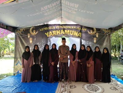 Nine individuals, one male and eight females, are positioned in a line under a temporary white canopy structure in Jambo Dalam, Indonesia. The male, wearing glasses, a brown patterned long-sleeved shirt, and a dark patterned sarong with a green emblem, stands slightly left of center. The eight females, appearing younger, are dressed in varying shades of long brown, maroon, or black dresses with black hijabs, standing to the male's left and right. Behind the group hangs a black banner with the gold and yellow text "LEMBAGA PENDIDIKAN ISLAM YARHAMUNA" and decorative crescent moon and lantern motifs. The ground immediately beneath the individuals is covered by a blue tarp on the left side and a patterned rug on the right. Beyond the banner and canopy perimeter, green foliage and trees are visible, with a portion of a motor vehicle partially observable on the far right. All individuals are standing still, facing the camera.