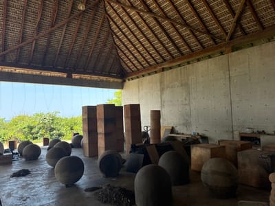 An interior wide-angle view depicts an open-sided workshop or storage facility located in San Isidro Llano Grande, Mexico, under bright daylight conditions. The structure features a prominent, high-pitched thatched roof supported by exposed wooden beams and a visible framework. The rear and right walls are constructed from unpainted, rough-textured concrete, displaying formwork tie holes. The left side of the building is open, presenting an unobstructed view of dense green foliage, with a distinct horizon line suggesting a body of water (likely the Pacific Ocean) beneath a clear blue sky. Several thin, dark wires or ropes hang vertically from the eaves on this open side.
The concrete floor within the facility is occupied by numerous large, roughly spherical or ovoid objects, appearing to be crafted from stone or concrete, scattered across the foreground and midground. Some of these objects exhibit variations in shape or apparent incompleteness. In the central and right areas of the space, several large cuboid objects, constructed from a reddish-brown material, are stacked in various configurations, forming tall columns or larger consolidated structures. To the far right, against the concrete wall, a workbench or shelf area is visible, holding various unidentifiable smaller items and what appears to be a hand truck or dolly near a stack of cylindrical objects. Irregular piles of dark, granular material (possibly dirt or debris) are distributed on the concrete floor. No individuals are present within the frame.