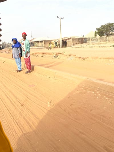 Two women stand on a sandy road in a sunny, rural environment. One wears a blue head covering, a striped shirt, and blue jeans. The other has on a red head covering, a light green shirt, and patterned red pants. Behind them is a scene of weathered buildings, fences, and power lines. Tire tracks are prominent in the sandy ground. The image has been rotated to the side.