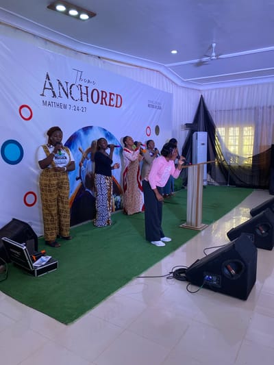 Here is a concise description of the scene:

The image captures a group of five women on a green stage in what appears to be a conference or event setting. The backdrop features a banner with the words "Theme ANCHORED, MATTHEW 7:24-27, YOUNG COUPLES RETREAT 2026", a circular design with images of people, and a series of red and blue circles. Each of the women is holding a microphone and appears to be singing or speaking into it. The woman nearest to the camera is wearing a patterned outfit with a shirt that reads "Voximi". The women are standing on a green carpeted stage. To the side of the stage are speakers and other sound equipment. The setting appears to be indoors, with white walls and ceilings. Based on the limited context, it's impossible to confirm the location within Miango, Nigeria, or any specific actions or interactions taking place at the event.