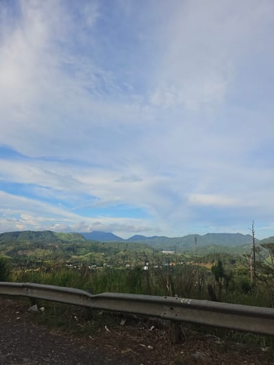 The image depicts a landscape scene featuring rolling green hills and mountains under a sky with scattered clouds. In the foreground, a gray metal guardrail runs horizontally across the frame, with a gravel road and dry grass visible below it. The guardrail shows signs of wear and has some white markings on it, possibly numbers or letters. Beyond the guardrail, the landscape transitions into lush vegetation, with trees and smaller plants growing densely. In the mid-ground, there are visible structures, suggesting human habitation or agricultural activity, including buildings and what appear to be cultivated fields or terraces on the hillsides. The distant background is dominated by layers of mountains fading into a hazy horizon. The overall impression is of a rural or mountainous region. The specific location in the city of Tu Póh, Vietnam, is not discernible from the visual elements alone.