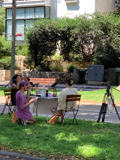 The image depicts an outdoor setting, likely in Tel Aviv, Israel, with lush green grass and trees surrounding a paved area. The time of day appears to be daytime, with bright sunlight casting shadows. The scene suggests a casual gathering or activity.
In the foreground, two individuals are seated. One person is wearing a purple t-shirt and appears to be holding a phone or similar object, looking downwards. The other individual is wearing a dark t-shirt and a purple head covering, also looking downwards, possibly engaged with the first person. Both are seated on black chairs.
Further back, another person is seated on a wooden chair, facing away from the camera. They are wearing a light-colored t-shirt with a graphic print. A white hairpin is visible in their dark hair. In front of this person is a table with various items, including what appear to be drinks (bottles and cups), books, and possibly some snacks.
Behind the seated individuals and the table, there is a stone wall and dense foliage from trees. In the background, there are several large, dark-colored industrial bins. To the left, a building with large windows and a balcony is visible, with more trees and greenery. A camera on a tripod is also present in the middle ground, suggesting some form of recording or filming might be taking place. The overall atmosphere seems relaxed and informal.