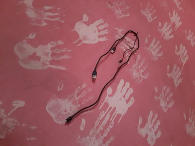 The image displays a pink wall adorned with numerous white handprints, some appearing faded and smudged, suggesting they were made with paint. Two black USB cables are laid out on the wall, crossing over the handprints. One cable has a USB-A connector at one end and what appears to be a USB-C connector at the other. The second cable, connected to the first, has a smaller connector, possibly a micro-USB or another USB-C. The arrangement of the cables suggests they might have been placed there intentionally, perhaps for display or as part of a creative setup. The handprints, varying in size and clarity, give the impression of a playful or commemorative scene, possibly related to children's artwork or a public event. The lighting suggests an indoor setting, and the time of day cannot be determined. No text is visible in the image. The scene is set in Uselu, Nigeria.
