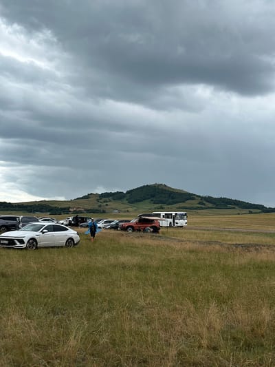 A wide-angle, eye-level, outdoor shot shows a parking lot filled with vehicles situated in a grassy field. In the foreground, a white sedan is parked. A person walks in the middle ground, carrying a blue object. A white bus and several other cars are parked further back. A small, tree-covered hill is visible in the distance, under a cloudy sky. The location is Bereznyakovka, Kazakhstan.