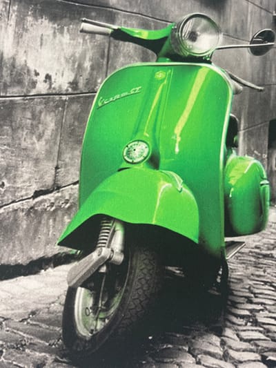 The media file shows a close-up, slightly angled view of a bright green vintage scooter parked on a cobblestone street next to a textured stone wall. The scooter is the primary subject, dominating the foreground and midground with its vibrant color. The background, rendered in black and white, depicts the rough texture of the stone wall and the patterned cobblestones of the street.
Notable details include the scooter's rounded bodywork, a prominent headlight, a handlebar with a mirror, and the rear wheel with its tire tread visible. A black shadow cast by the scooter falls across the stone wall. The image has a slightly grainy texture, suggesting it might be a photograph or a print with a textured surface.
There is no visible text in the media file. The scene appears to be set outdoors during the daytime, with the lighting suggesting clear weather. The overall impression is one of a static display, with the scooter simply parked and observed. The location context is Calp, Spain.