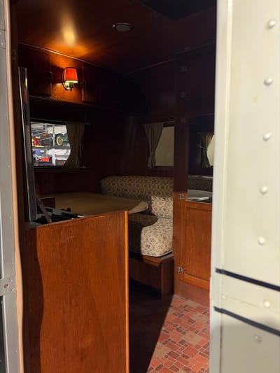 This image captures the interior of what appears to be a vintage cabin or compartment, possibly within a vehicle like a bus, train, or RV, likely displayed in a museum setting in Wassenaar, Netherlands. The perspective is tilted, but the scene primarily features warm, dark wooden paneling on the walls and furniture.
A light-colored, possibly white, ceiling or upper structure is visible at the top, featuring several small, round rivets or bolts. Below this, a series of wooden shelves or compartments are built into the wall. On the right side, a seat or cushion upholstered in a patterned fabric with floral or botanical motifs is prominent, alongside another padded surface, possibly a bunk or storage area, above it. Further to the right, a wall or floor section displays a distinctive brick-like pattern in shades of brown and orange.
On the left side, a vintage wall light fixture with an orange-tinted shade emits a warm, soft glow onto the wooden wall, creating an inviting atmosphere. Below the light, a rectangular window or opening offers a glimpse, possibly a reflection, of an external scene that includes mechanical parts and a yellow wheel, further suggesting an exhibition environment.
No people are present, and there is no visible text. The interior design emphasizes historical craftsmanship and a cozy ambiance, likely enhanced by the warm artificial lighting.
