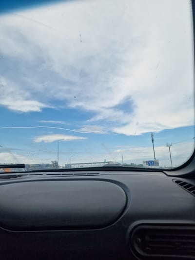 The image is taken from inside a vehicle looking out through the windshield. The view shows a bright blue sky with scattered white clouds. A thin, white horizontal line, possibly a contrail from an airplane, is visible across the sky. In the distance, there are road signs, streetlights, and cellular towers. The dashboard of the vehicle is visible in the foreground, appearing dark and textured. The overall scene suggests a daytime drive on a highway or a similar road. There is no specific indication of the location being Zmeika, Russia, or any other city.