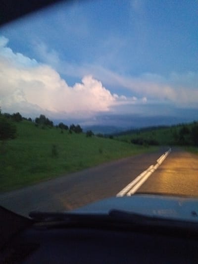 The image depicts a view from a vehicle driving on a paved road, likely at dusk or early evening. The road has white dashed lines marking the lanes. To the left of the road is a grassy hillside dotted with trees. In the distance, rolling hills are visible under a sky with scattered clouds, some of which are illuminated by soft pink and white light. The front of the vehicle's dashboard is visible in the foreground. The location is identified as Sadovyi, Russia.