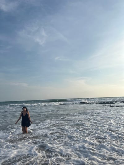 The image shows a woman standing in the shallow water of a beach, located at the coordinates 9.635 latitude and -85.161 longitude in Santa Teresa, Costa Rica. The woman is wearing a black dress. The beach is sandy and covered in white foam from the waves. The ocean is rough with large waves breaking in the distance. The sky is light blue with white clouds. The scene is bathed in the light of a setting sun. The woman is facing the camera and smiling. Her hair is long and dark. The image was taken at eye level and in landscape orientation.