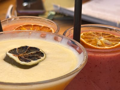 The image presents a close-up view of three cocktails on what appears to be a wooden surface. The foreground cocktail is a light-yellow color with a dried lime slice as a garnish. Behind it, a cocktail with an orange hue is garnished with a dried orange slice. To the right, a reddish-pink cocktail also has a dried orange slice on top. Black straws are visible in the yellow and reddish-pink cocktails. In the background, there are blurred objects including a pair of glasses and papers. The setting appears to be a restaurant or bar setting, however, there is no available information about the location being in Cala D'or, Spain.
