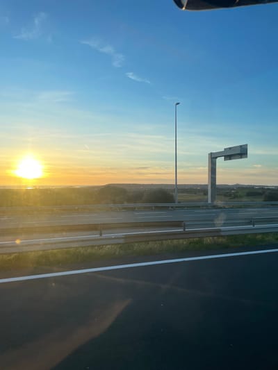 The image displays a highway scene at either sunset or sunrise, located near La Palme, France. The view is from a vehicle, with the upper edge of its interior visible at the top. The foreground features dark asphalt road with a thick white line marking the lane edge. Above this, a two-bar metallic guardrail runs horizontally across the frame. Beyond the guardrail, multiple lanes of a paved highway are visible, delineated by white markings.
The horizon on the left is dominated by a bright, intense sun, casting a golden-orange glow that reflects strongly off a distant body of water, indicative of the Étang de La Palme. Above the highway and further into the midground, a tall, grey streetlight pole stands vertically. To its right, a large, grey gantry structure for an overhead road sign is present, with its sign face appearing blank. The background beyond the highway consists of a flat to gently undulating landscape covered with scattered green vegetation and leading to low hills in the distance. The sky transitions from a brilliant golden-orange near the horizon to a clear light blue overhead, featuring a few wispy white clouds and a faint aircraft contrail. No people or active interactions are visible.