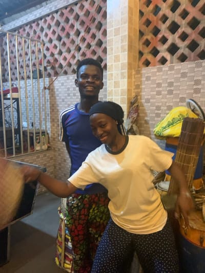 This image captures two young men standing and smiling, likely indoors, in what appears to be an evening setting. The overall mood conveyed is positive and friendly.
The man in the foreground, positioned slightly to the right, is wearing a white short-sleeved t-shirt with a black trim around the collar. He has a black head covering, possibly a durag or bandana. He is smiling broadly and looking slightly to his left (our right). His lower body is visible, showing dark blue trousers with small, light-colored polka dots or patterns. He is holding a distinctive musical instrument in his right hand. The instrument is long and cylindrical, composed of numerous thin, parallel wooden sticks or reeds bound together, suggesting a traditional percussion or shaker instrument. His left arm is bent, resting near his waist.
Behind him, slightly to the left, stands another young man. He is wearing a blue short-sleeved t-shirt with white stripes along the sleeves. He is also smiling and looking in a similar direction as the first man. Both men appear to be of African descent.
The background features patterned tiled walls. On the left side, behind the man in blue, the wall has a reddish-brown, diamond-grid pattern on a lighter base. The wall section behind the man in white shows a lighter, possibly cream or off-white, tiled pattern with smaller, repeating geometric shapes.
To the left of the man in blue, there is a metallic structure resembling a multi-tiered rack or shelf, with several horizontal bars. Behind these bars, some dark shapes and small indicator lights are vaguely visible, hinting at electronic equipment, possibly related to audio or music production.
In the lower right corner, and also behind the man in white, various colorful fabrics with intricate patterns are visible, suggesting traditional African textiles or household items. There is also a yellow bag and what looks like a part of a bicycle wheel further in the background, near the top right when the image is viewed upright.
The lighting in the image is somewhat dim but focused, suggesting artificial light sources illuminating the subjects against a darker background, reinforcing the impression of an indoor or covered evening setting. No text is visible in the image.