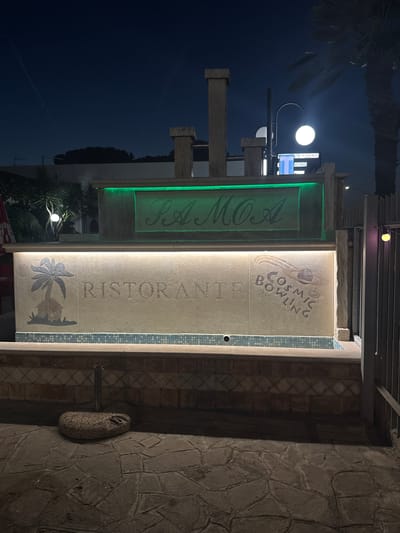 The image depicts a nighttime view of a building facade in Ardea, Italy, specifically at "Cosmic Bowling". The facade features a sign with green neon lights that reads "RAMOA". Below this, a beige wall is illuminated by warm lights. On this wall, a stylized palm tree and hut is etched on the left, and the word "RİSTORANTE" is carved in the center. To the right, a graphic depicts a bowling ball with swirling patterns, and the words "COSMIC BOWLING" are etched in a circular fashion. A tiled border runs along the bottom of the beige wall. The foreground shows a stone-paved walkway with a large, dark stone base for an umbrella or similar object. The background features a dark sky with a few faint contrails, trees, and streetlights. The overall scene suggests an entertainment venue or restaurant open at night.