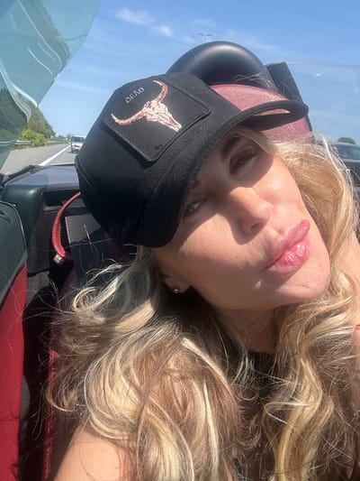 A woman with blonde, wavy hair is the focal point of the image, possibly in Leidschendam, Netherlands, though specific city landmarks are absent from the frame. She is wearing a black hat with a steer skull logo. The environment suggests she is in a vehicle with red leather seats and a windshield. The lighting is bright, indicating daytime.