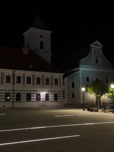 The image displays a night scene of an urban architectural setting in Osijek, Croatia, characterized by historic buildings and modern ground illumination.
Two prominent buildings anchor the background. On the left, a multi-story edifice features a grey facade, a dark, angled roof, and a distinct tower-like structure with small windows near its peak. Its lower sections are adorned with numerous rectangular windows, some of which have dark grates or balustrades. To its right stands another building, also with a grey facade, showcasing a blend of arched and rectangular windows, and decorative cornices. Several traditional-style streetlights are mounted on this building's facade, illuminating parts of it and some green foliage growing nearby.
In the foreground and extending towards the right, a wide, light-colored paved area, likely a pedestrian square or street, dominates. This surface is uniquely illuminated by multiple parallel, glowing white light strips embedded directly into the ground, creating a striking linear pattern across the scene.
The time of day is clearly night, with a completely black sky and all visible light originating from artificial sources: streetlights, internal window lights, and the distinctive ground lighting. The scene is devoid of people or discernible activity, conveying a quiet and tranquil atmosphere. No visible text is present in the image. The architecture suggests a European historical context, aligning with the provided location of Osijek, Croatia.