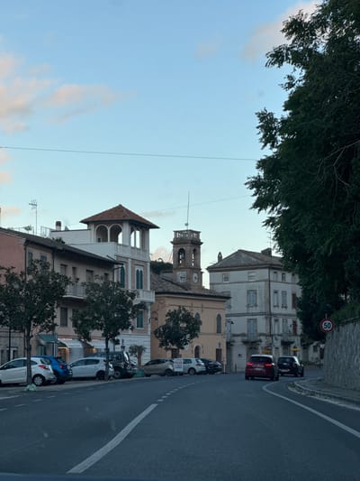 The image displays a street view in Pedaso, Italy, characterized by historic architecture and vehicles. The perspective is from within a vehicle, looking forward along an asphalt road with white lane markings.

On the left side of the road, a series of multi-story buildings stand, featuring various facades and window styles. One prominent light-colored building has a distinctive multi-arched, terracotta-tiled belvedere on its roof, along with balconies and green window shutters. Adjacent to it, a clock tower with visible bells and a terracotta roof rises, indicative of a church or public building. Further right, additional multi-story structures with faded, lighter-colored facades are visible.

A row of parked vehicles lines the left side of the street, including several hatchbacks (white, blue, grey) and a dark SUV. One white parked car displays a "PIZZA EXPRESS" sign on its side. On the roadway, two vehicles are actively moving away from the viewer: a red hatchback or compact SUV leads, followed by a dark-colored SUV or sedan.

The right side of the road features a stone wall and a large, dense tree canopy that partially obscures the sky and background buildings. A circular speed limit sign indicating "50" is affixed to the right wall. The sky above is light blue with scattered, wispy clouds. Power lines traverse the upper portion of the scene. No individuals are visible.
