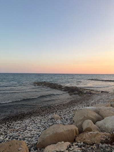 The image depicts a tranquil beach scene in Pervolia, Cyprus, at dusk. The sky transitions from a clear pale blue at the top to a soft gradient of pink and orange near the horizon, indicating the setting sun. The sea is a deep blue-green, with gentle waves lapping at the shore. A man-made breakwater, constructed from large, irregular stones, extends into the water, creating a sheltered cove. The beach itself is covered in a mixture of small white and grey pebbles and larger, tan-colored rocks. The foreground is dominated by several large boulders, one of which is partially covered in dry, brown vegetation. The overall impression is one of peaceful natural beauty.