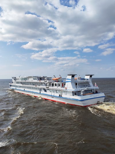 A wide-angle, eye-level outdoor shot captures a large white passenger ship with blue and red accents sailing on a choppy, dark brown body of water. The ship, identified by Cyrillic lettering on its stern as "СЕРГЕЙ ЕСЕНИН" and "МОСКВА," is oriented at a slight angle, moving from the left of the frame towards the right. The vessel features multiple decks with numerous windows and railings, life preservers, and lifeboats visible. Two dark smokestacks are prominent on the upper rear deck. A Russian flag is affixed to the stern. A small group of individuals, appearing as silhouettes, are visible on the deck. The water is disturbed, with white foam and wakes trailing from the ship's stern. The upper portion of the image is dominated by a bright blue sky with scattered, fluffy white clouds. The scene is likely set on a river or large lake, given the presence of a passenger ship and the calm but wavy water. The location in the city of Krokhino, Russia is not visually discernible from the image itself.