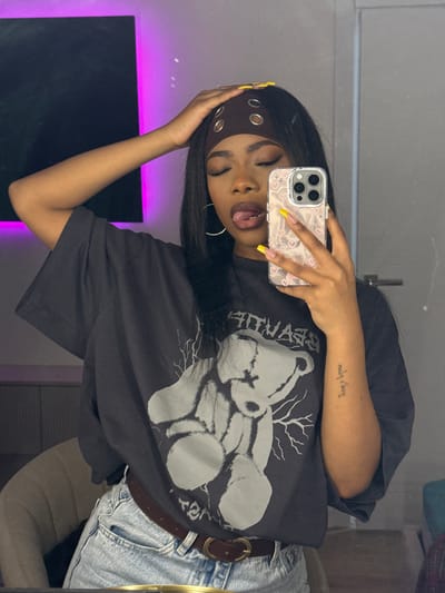 A person is taking a selfie in what appears to be an indoor environment. The person is wearing a dark-colored t-shirt with a graphic of a teddy bear, a brown headband with metal eyelets, and light-wash denim jeans with a brown belt. They are holding a cell phone with a light-colored case, showing a yellow-tipped nail on their finger. The person has dark hair and is sticking their tongue out. There is a tattoo on the person's left arm. A dark-colored object, possibly a television, is visible in the background with a purple light source illuminating the wall behind it. The location, Troitsk, Russia, is not discernible from the provided information.