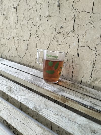 A close-up, eye-level shot shows a glass mug filled with amber-colored liquid, resting on weathered wooden planks. The mug, partially obscured by the foreground planks, features green text that reads "Alokozay PEPPERMINT" and a decorative green leaf motif. The background is a textured, cracked earth wall, suggesting a dry environment. The scene is set outdoors in Aktam (Tashminvody), Uzbekistan, evident from the typical architecture and natural elements of the region. No people are present in the image.