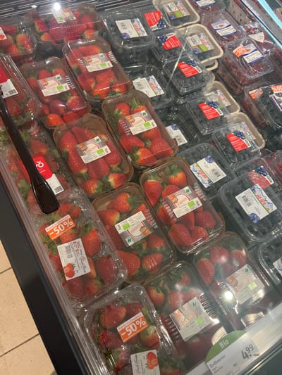 This media file captures a close-up view of a refrigerated produce display in a supermarket located in Bamberg, Germany. The primary subjects are numerous plastic containers filled with fresh berries, mainly strawberries and blueberries.
The strawberries are packaged in clear plastic punnets, many of which carry labels indicating "ERDBEEREN" (strawberries) and a weight of "300g." Several of these labels also feature a "BIO" (organic) certification logo. A prominent detail is the presence of red and orange stickers on some strawberry containers, advertising "Kurze Haltbarkeit!" (short shelf life!) and a "50%" discount. Brand names such as "HEIDL" and "Fresh Farm" are visible on the packaging.
Blueberries are also displayed in clear plastic containers, with some appearing to have a molded fiber base inside, also bearing "HEIDL" and "BIO" labels. In the upper right section of the image, a larger white price tag is visible, indicating "Bio ERDBEEREN" in a "300g Pack" for "4.99," with a smaller number "6.63" likely representing a comparison price. The items are neatly arranged and stacked in the brightly lit display case, with reflections visible on the plastic surfaces. The scene depicts a typical retail environment for fresh produce, implying an activity of grocery shopping. No people are visible in the image.