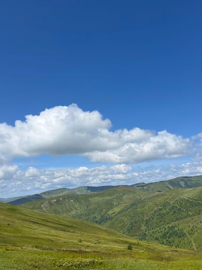 The image depicts a vast, undulating landscape of green hills under a bright blue sky dotted with fluffy white clouds. The terrain is covered in lush grass and scattered trees, with visible winding paths traversing the slopes in the distance. The scene appears to be a natural environment, likely a mountain range, in the vicinity of Chorna Tysa, Ukraine. No people, objects, or actions are discernible in the image, focusing solely on the expansive natural scenery.